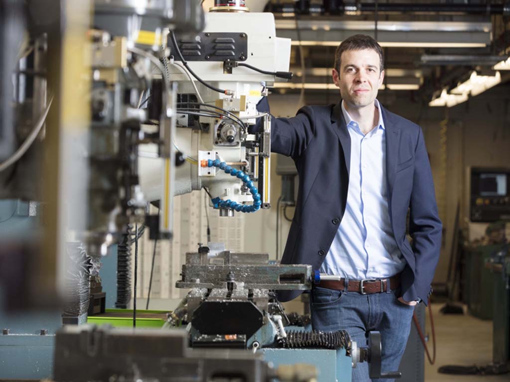 John Hart stands next to 3D printing hardware in a laboratory