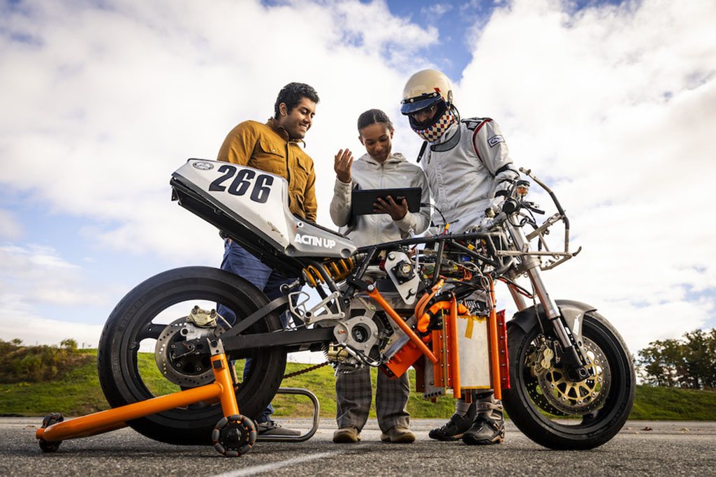 Three people, one wearing a crash helmet, check data on a screen attached to a motrocycle. 