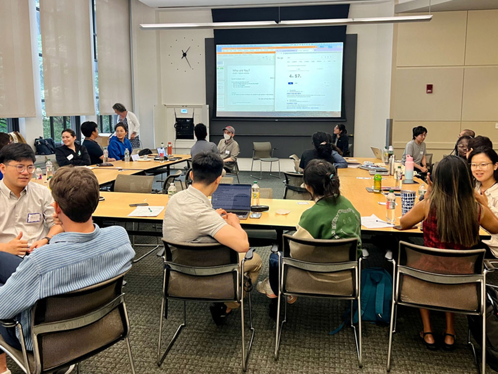 A meeting room is filled with about 20 students, seated, in one-on-one conversations with each other. A projection screen in the background shows a document with the title “Who Are You?”