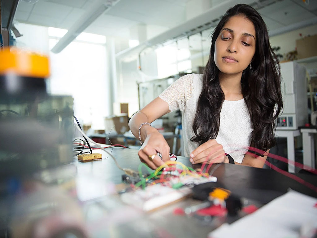 Ritu Raman working at a table in a lab, she is wearing a white shirt, and looking down towards her materials.