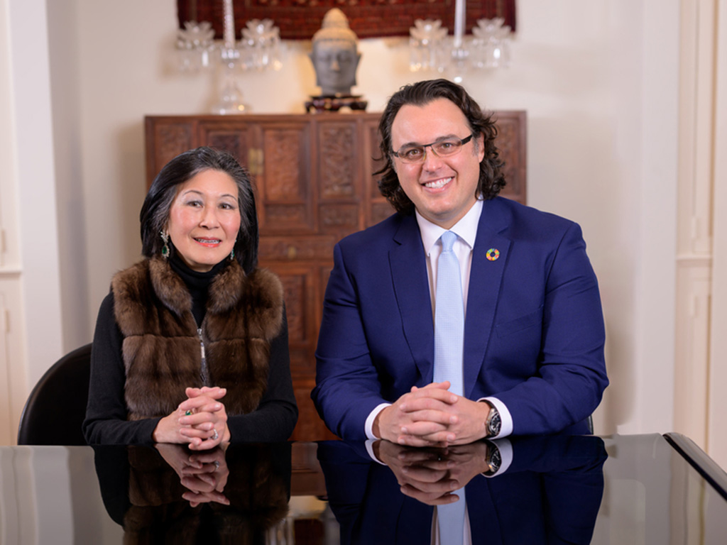 Lisa Yang and Amos Winter pose for a photo. They sit together at a black table in a white room with an intricately carved wooden dresser and a Buddah head statue in the background.